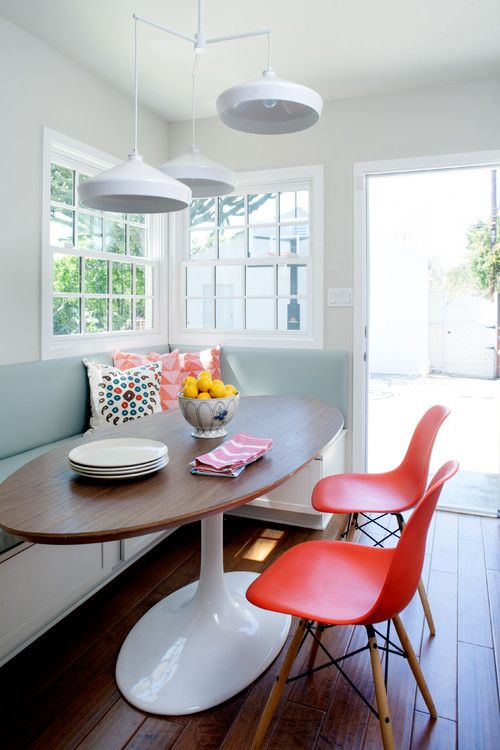 a mid-century modern dining space with a light blue upholstered bench, an oval table, red Eames chairs and white pendant lamps