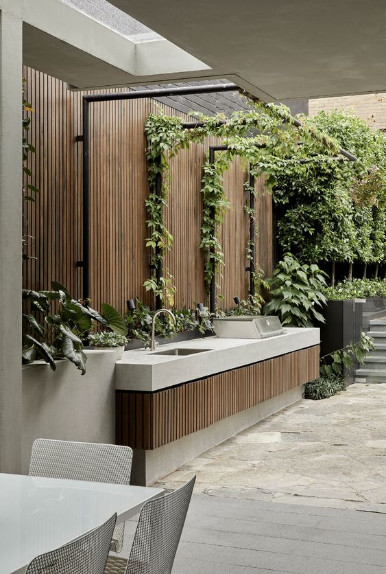 a minimalist bbq zone with a concrete kitchen island with fluted wood decor, a bbq and a sink, some greenery and a roof over the space