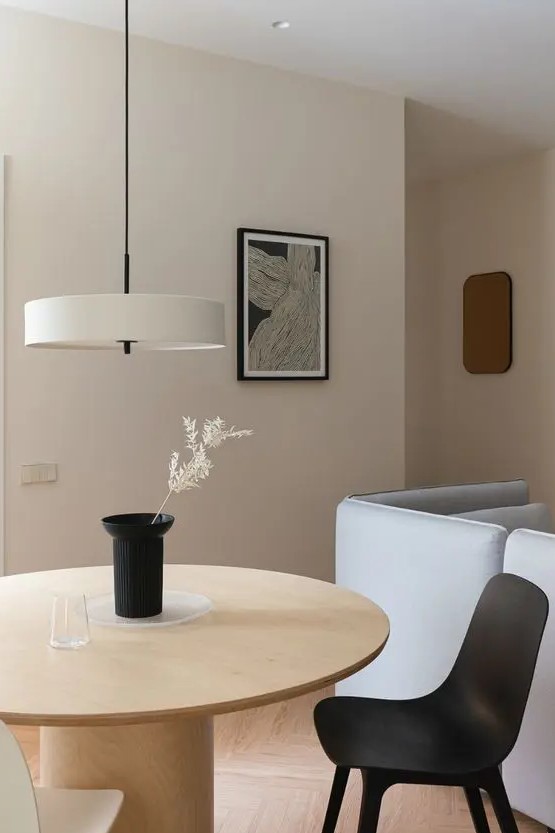 a minimalist dining space with a round light-stained table, a black and neutral chair, a neutral pendant lamp and a black vase