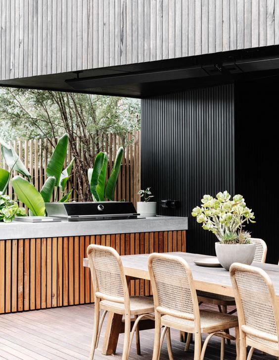 a minimalist outdoor bbq area with a concrete cabinet holding a grill, a stained table and wicker chairs and some greenery