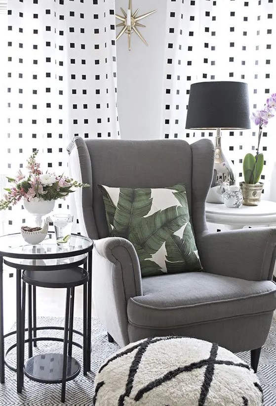a modern cozy nook with a grey Strandmon chair, glass side tables, a printed pouf, a table lamp and printed curtains