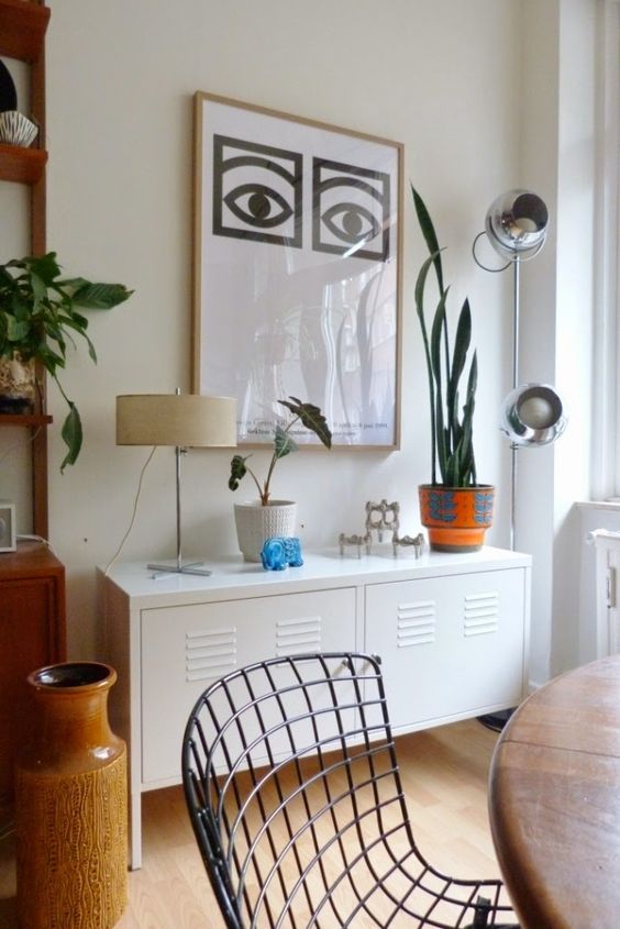 a modern living room with a white locker console, a stained table and metal chairs, a bold artwork, potted plants and lamps