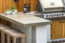 a modern outdoor bbq area with a large tiled unit with a grill, a concrete countertop, wooden stools and potted greenery