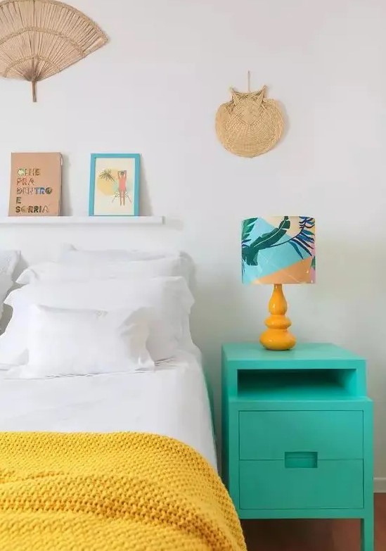 a modern tropical bedroom with a bed with yellow and white bedding, with a turquoise nightstand, a bright lamp and some woven touches to decor