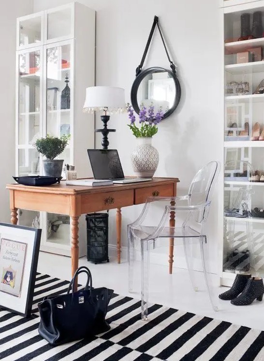a monochromatic home office with glass storage units, a stained vintage desk, a ghost chair, a striped rug and a round mirror