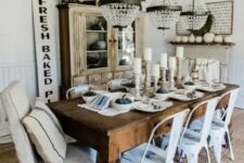 a neutral farmhouse dining room with a shabby chic buffet, a dark-stained table, white metal chairs, a ladder with crystal chandeliers