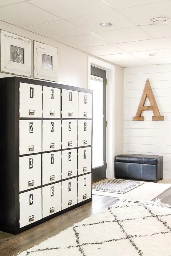 a neutral farmhouse mudroom with white beadboard walls, a large locker unit with numbers and a black leather upholstered bench