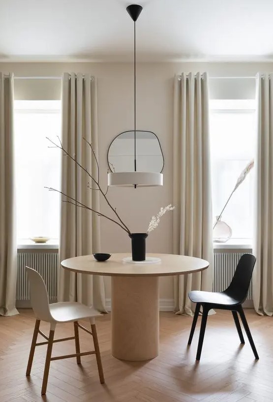 a neutral minimalist dining space with a round table, a black and a neutral chair, a creamy pendant lamp and a vase