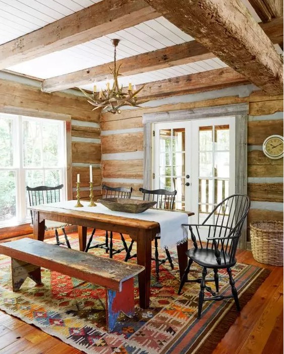 a rustic meets chalet dining room with stained wood on the walls and ceiling, a stained table and benches, black chairs and an antler chandelier