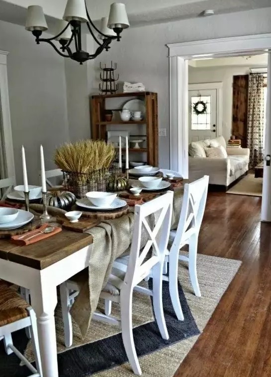 a rustic vintage dining space with grey walls, a white table with a stained tabletop, white chairs, a vintage chandelier and wheat centerpiece plus some candles