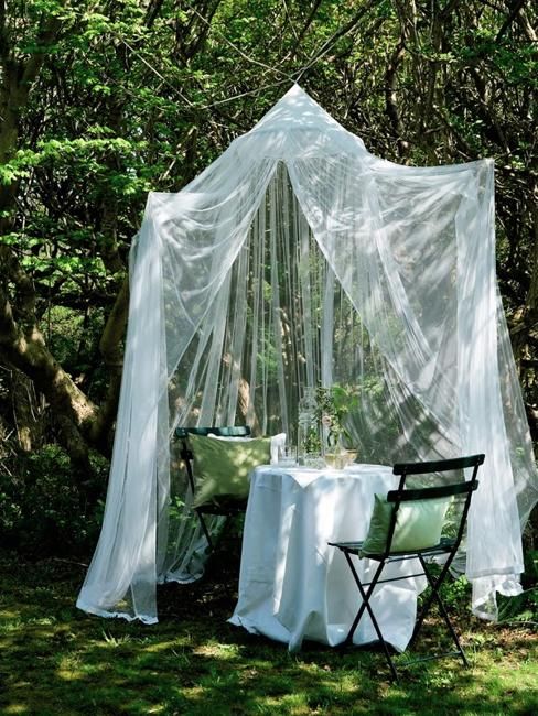 a small and cool outdoor dining space with a table, some chairs, a neutral mosquito net canopy and some greenery