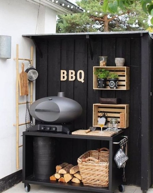 a small and cute outdoor kitchen of a black grill table with a basket and firewood, crate shelves attached to the wall and greenery