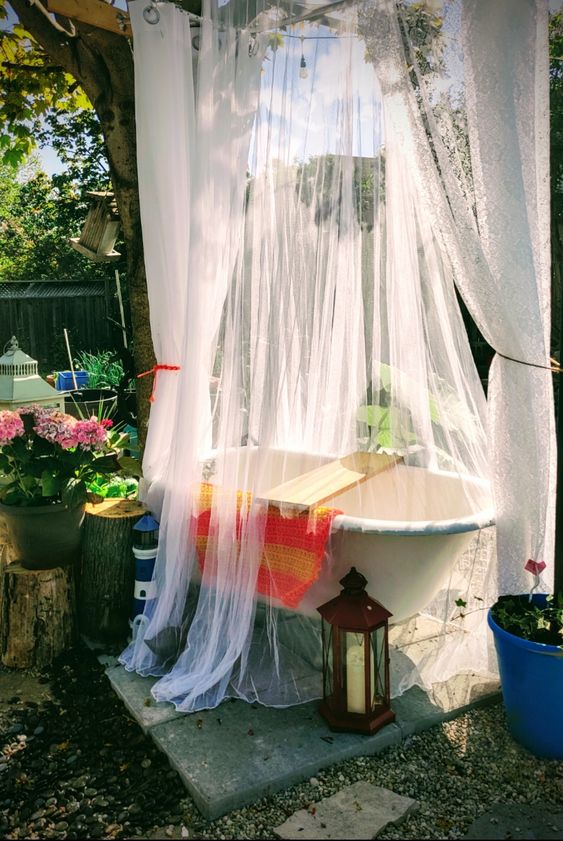 a small and lovely outdoor bathroom with a bathtub on a stand, with a mosquito net canopy over the tub, potted blooms and a lantern