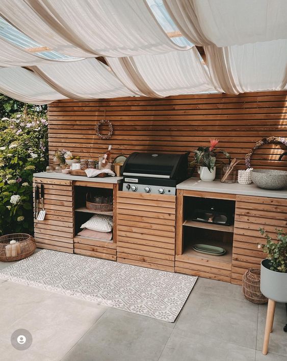 a small and modern outdoor bbq area with a large wooden unit, a grill, storage shelves and some potted blooms and herbs