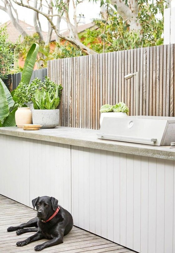 a small and sleek contemporary outdoor kitchen with a white planked cabinet and a concrete countertop, a grill, potted plants is very elegant