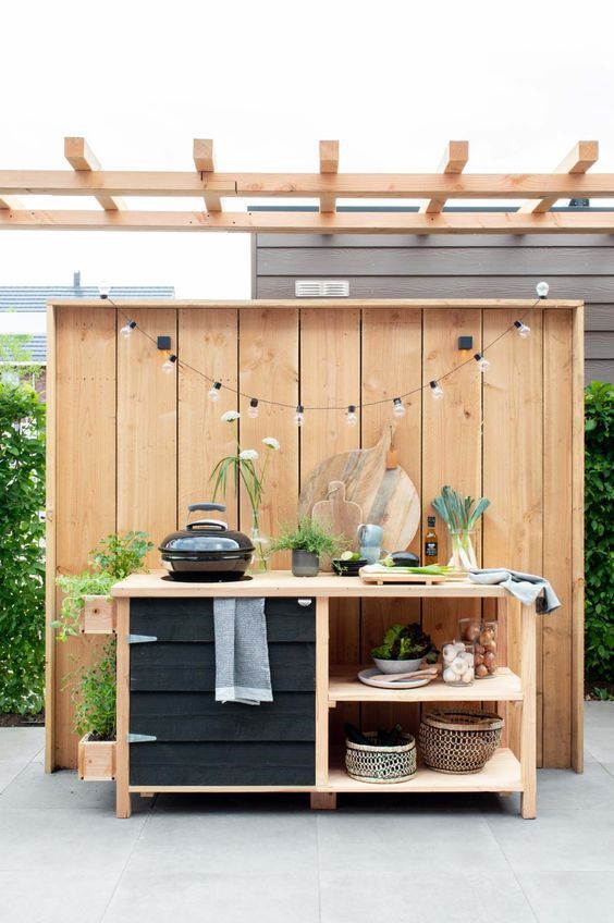 a small farmhouse outdoor grill table with shelves under a pergola, a grill and some herbs and greenery is a cool idea