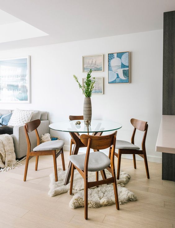 a small modern dining zone with a glass dining table with a wooden base, a rug and light-stained chairs plus a gallery wall