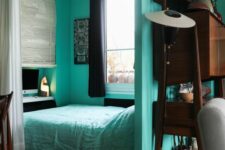 a small modern sleeping space with turquoise walls and a bed with turquoise bedding, a desk and a chair and black curtains