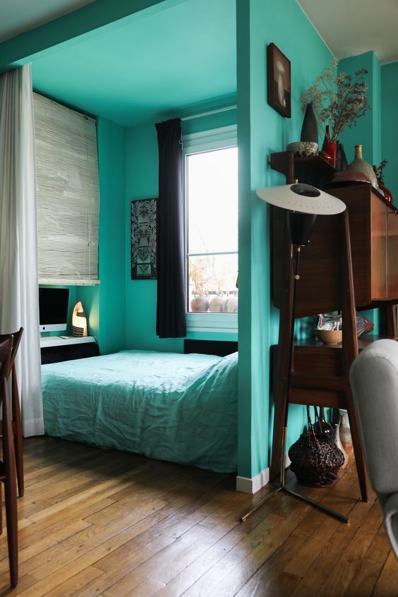 a small modern sleeping space with turquoise walls and a bed with turquoise bedding, a desk and a chair and black curtains