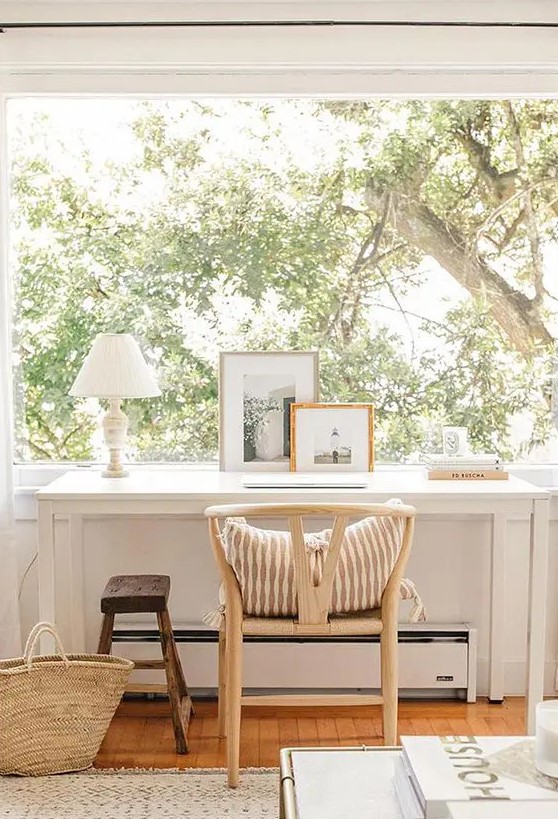 a small working nook by the window, with a sleek white desk, a wishbone chair and a small stool, a lovely greenery view