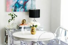 a small yet chic dining space by the window, with a round table, ghost chairs, a mirror console table, a bold artwork and a crystal chandelier