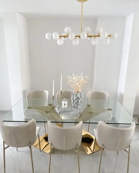 a sophisticated dining room with a large polished gold base and a glass tabletop, creamy chairs and a mid-century modern chandelier