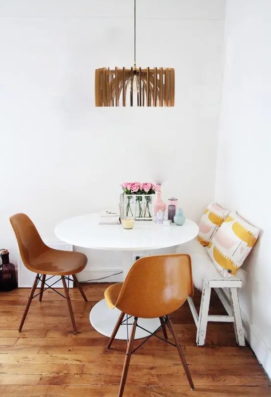 a stylish dining nook with a white bench and printed pillows, a round table, amber Eames chairs and a wooden lampshade