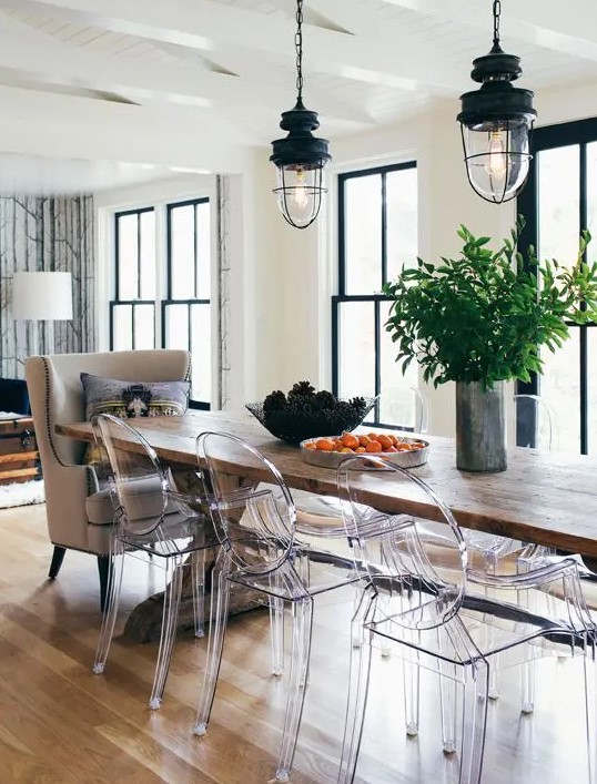 a stylish eclectic dining space with a long stained table, a neutral chair and some ghost ones, black vintage lamps on chain