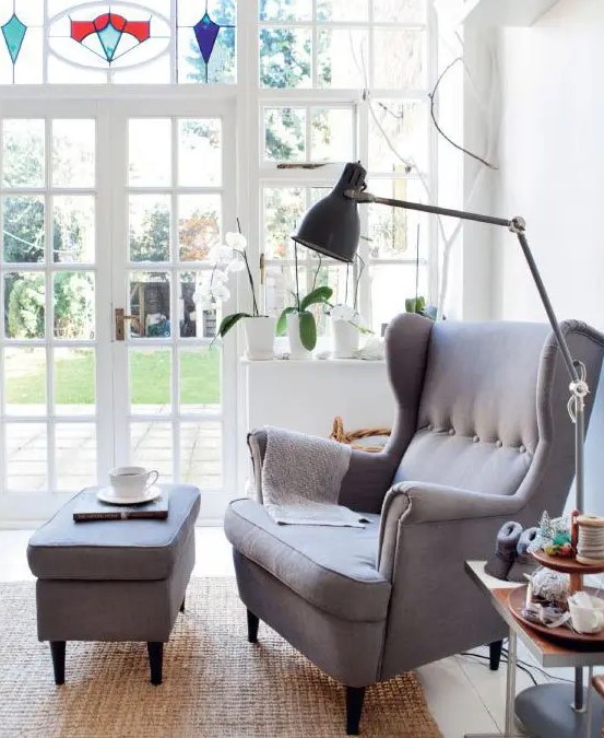a stylish reading nook by the glass doors, with a grey Strandmon chair and ottoman, a black lamp and a side table