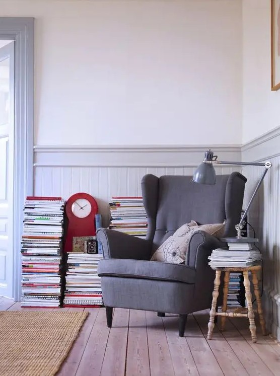 a vintage reading nook with a grey Strandmon chair, a wooden stool, stacked books and magazines and a grey floor lamp