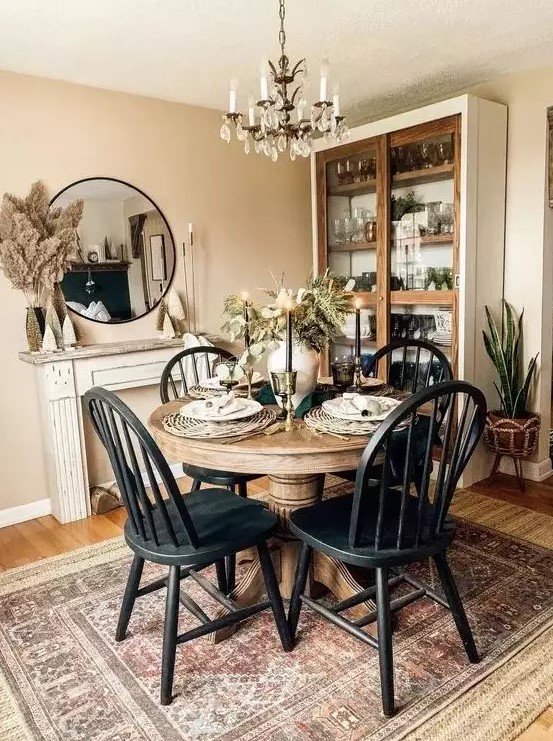 a vintage rustic dining space with a staiend round table, matching black chairs, a crystal chandelier, a glass buffet and a faux fireplace with a round mirror over the mantel