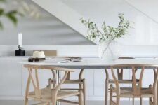 an airy Scandinavian dining space with a white dining table, light-stained wishbone chairs, a sleek storage unit and greenery