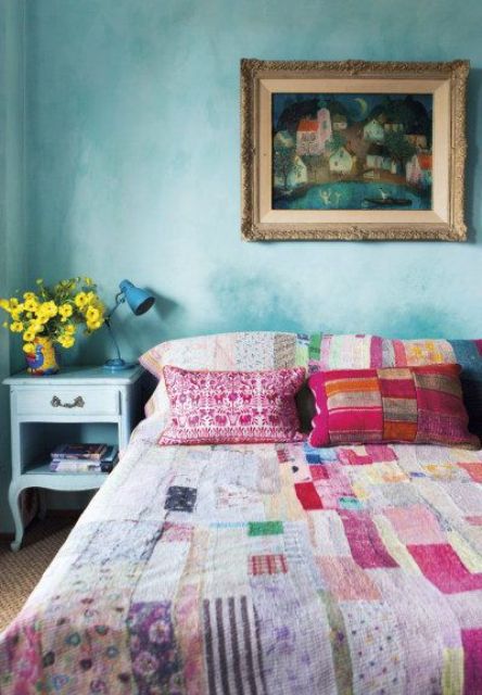 an eclectic bedroom with turquoise textured walls, a bed with bright bedding, a light blue nightstand and a bold artwork
