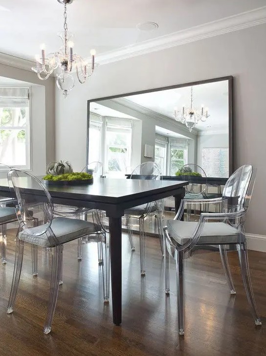 an elegant dining room with a black dining table, ghost chairs with grey cushions, a chic chandelier, an oversized mirror
