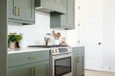 an elegant olive green kitchen with shaker cabinets, gold and brass handles, a white tile backsplash and a printed rug is cool