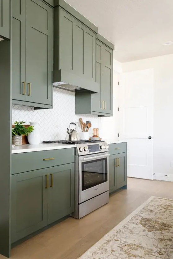 an elegant olive green kitchen with shaker cabinets, gold and brass handles, a white tile backsplash and a printed rug is cool