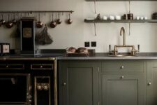 an elegant vintage olive green kitchen with shaker cabinets, grey stone countertops, a large hood, copper and brass touches