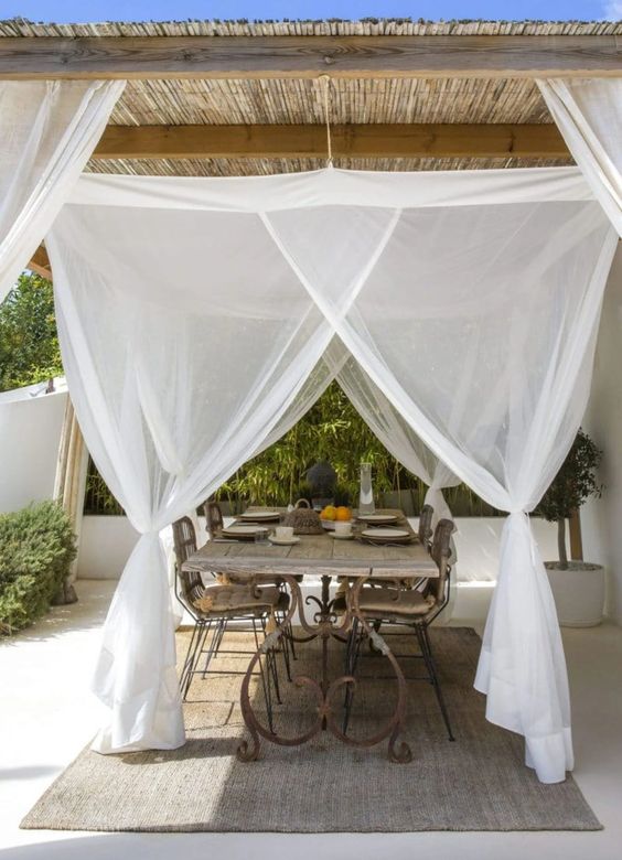 an outdoor dining space with a vintage rustic table and wicker chairs, a mosquito net canopy and some greenery