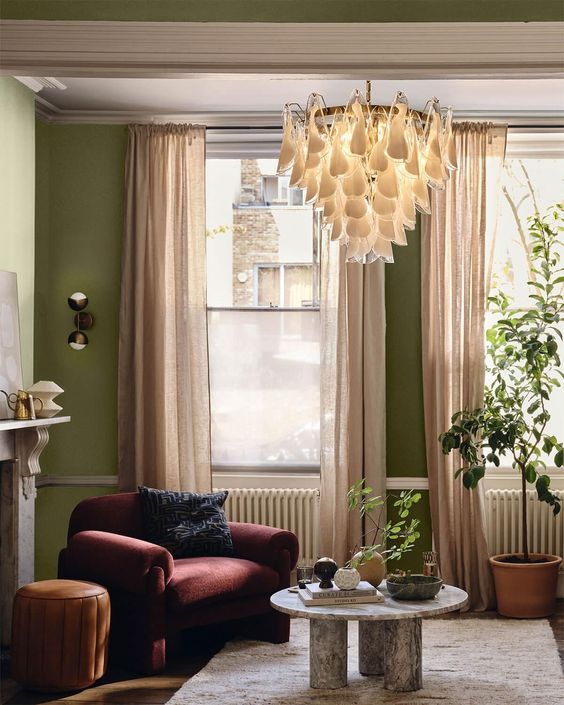 a bright living room with olive green walls, a burgundy chair and an orange pouf, a chandelier and blush curtains
