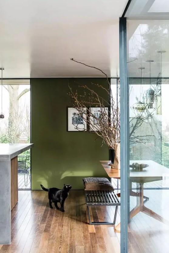 a cool dining space with an olive green accent wall, a wooden trestle dining table, a wooden bench and some branches in a vase