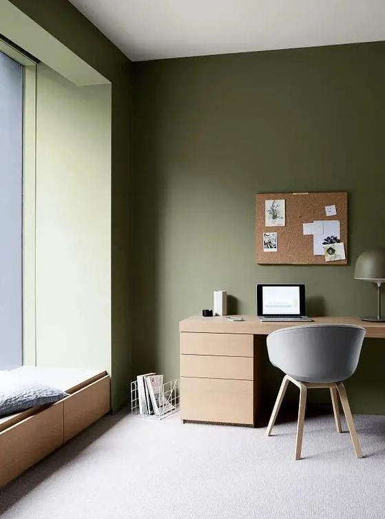 a minimalist olive green home office with a windowsill daybed, a stained desk and a grey chair, a cork board and a lamp