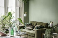 15 a refined living room with an olive green wall, a moody green sofa, some glass tables and lots of greenery