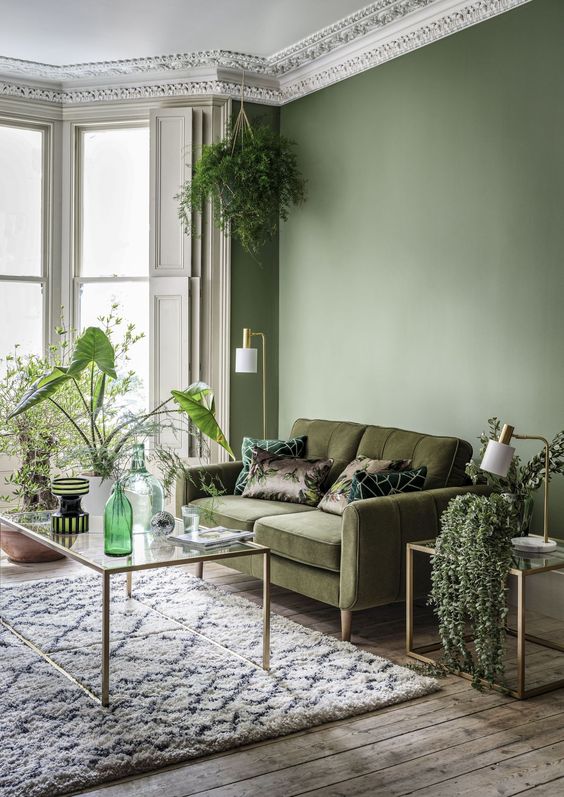 a refined living room with an olive green wall, a moody green sofa, some glass tables and lots of greenery