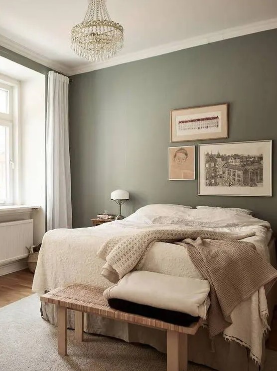 a relaxing bedroom with olive green walls, a bed with neutral layered bedding, a woven bench, a crystal chandelier and some art