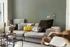 19 a Scandinavian living room with olive green walls, a grey sofa and colorful pillows, a gold sconce, a rocker and some coffee tables