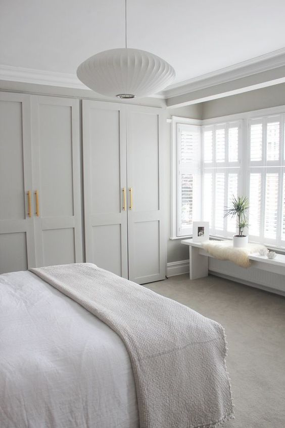 a neutral bedroom with dove grey IKEA Pax wardrobes with gold handles, a large bed with neutral bedding, a bench with cool decor