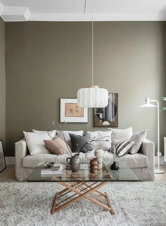 an airy living room with olive green walls, a neutral sofa and pillows, a glass coffee table, a pendant lamp and some art