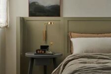 26 an elegant bedroom with olive green paneling, a bed with olive green and neutral bedding, a grey nightstand and a gold sconce