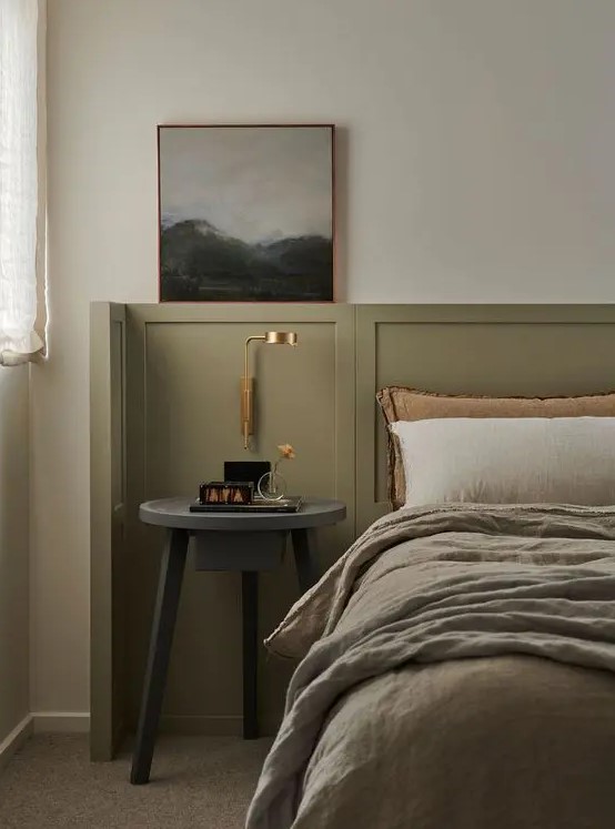an elegant bedroom with olive green paneling, a bed with olive green and neutral bedding, a grey nightstand and a gold sconce