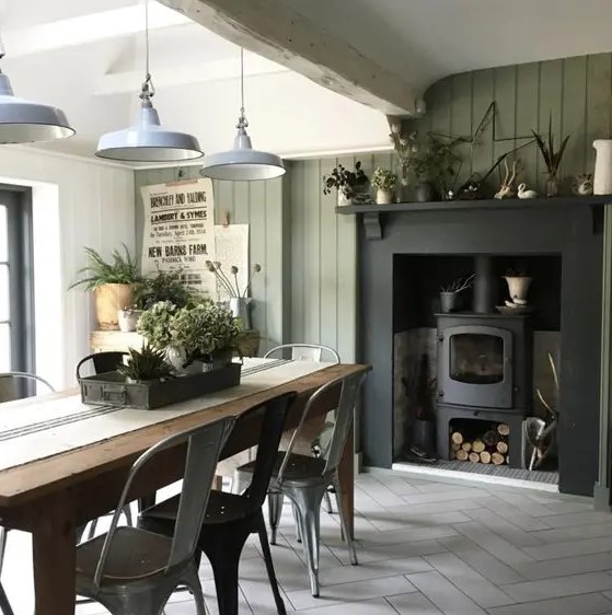 an industrial farmhouse dining room with an olive green shiplap wall, a hearth, a wooden table and metal chairs, metal pendant lamps